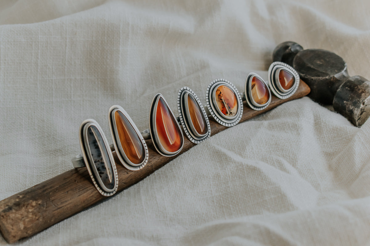 Seven sterling silver rings sit atop an antique hammer. Each ring is made from silver and Montana Agate stone, and are unique in shape, size, and design.