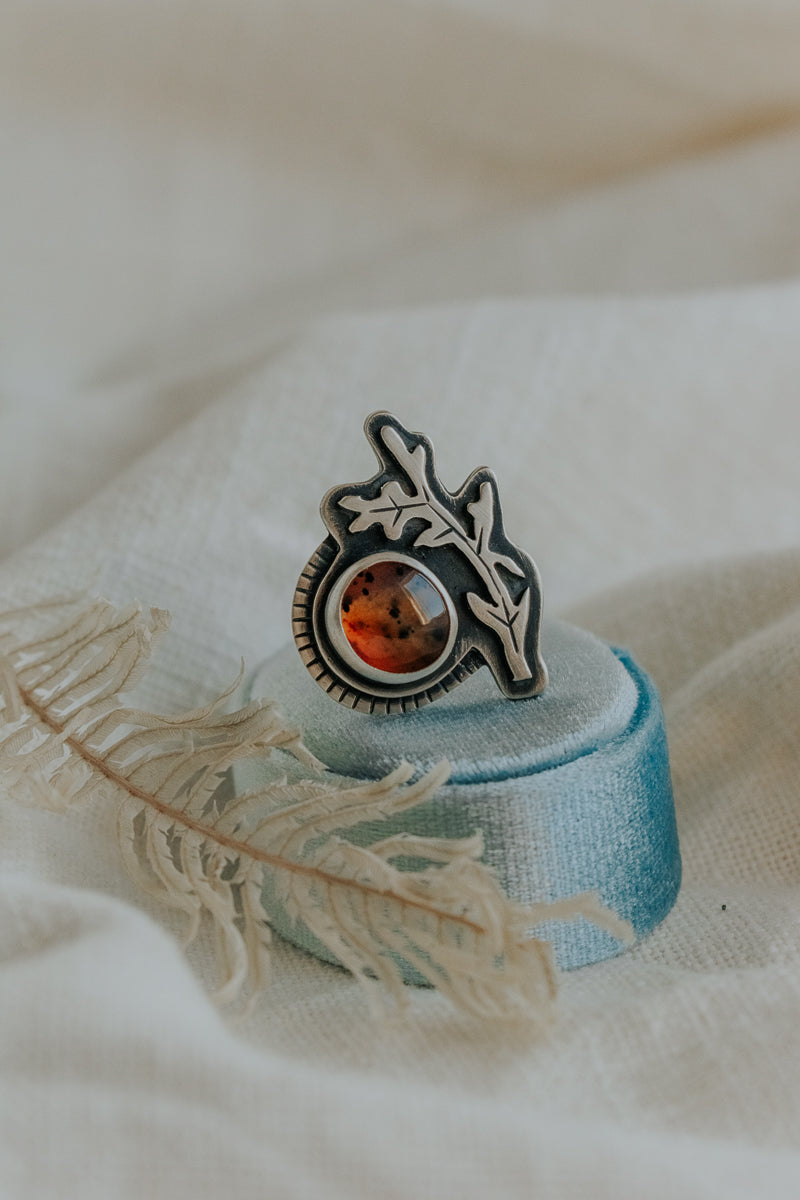 A ring made from a round Montana Agate stone with a sterling silver leaf or branch hugging around the stone.  The ring is sitting on a light blue velvet ring box against a white cloth backdrop.