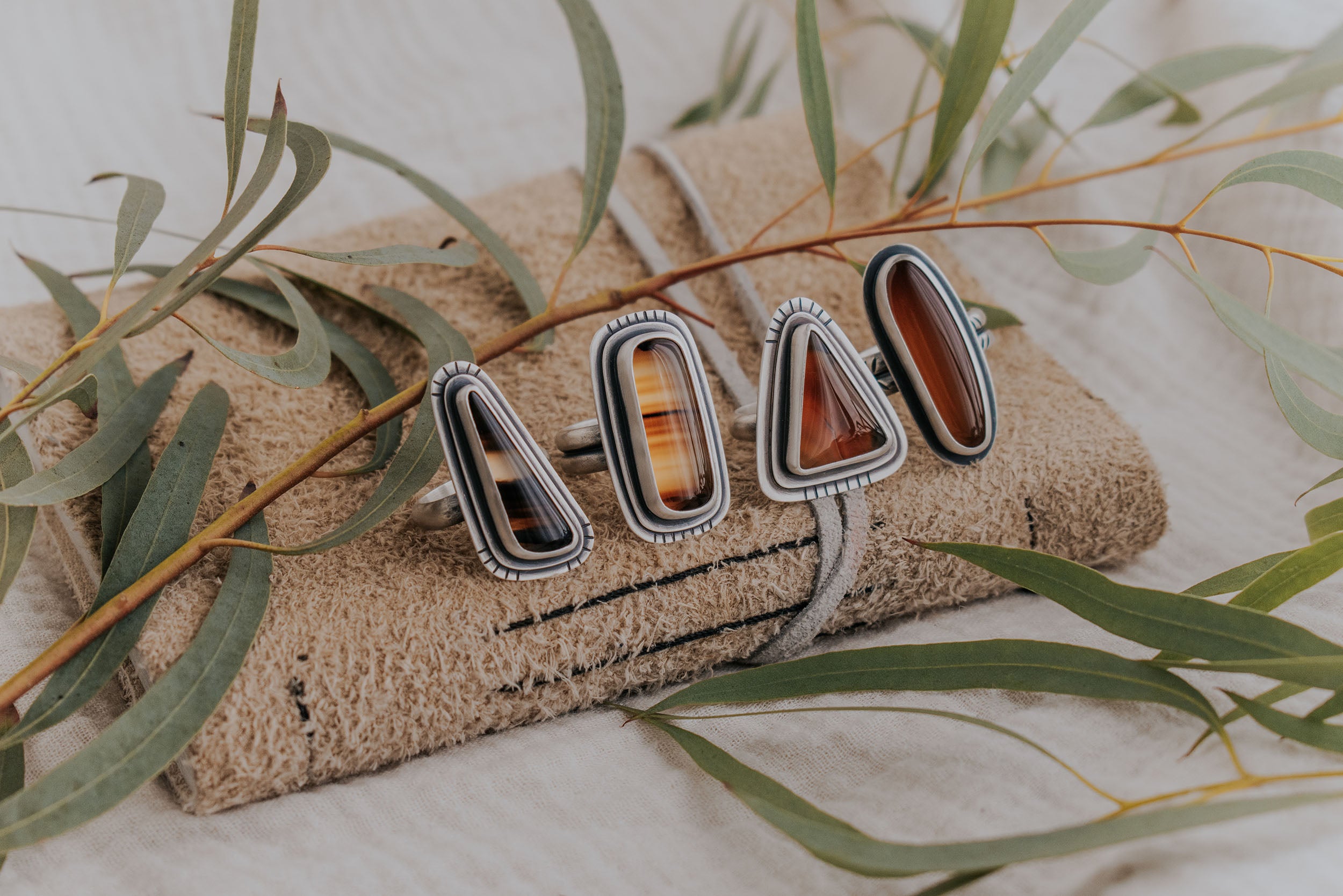 A set of four Montana Agate rings sit on a leatherbound notebook surrounded by green dried foliage. 