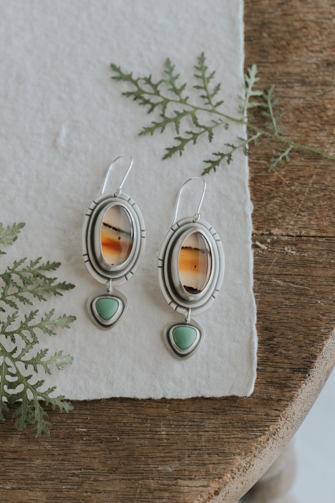 A pair of earrings is pictured against a backdrop of wood and leaves. The earrings are made from sterling silver, oval Montana Agate stones, and smaller triangular variscite stones. 