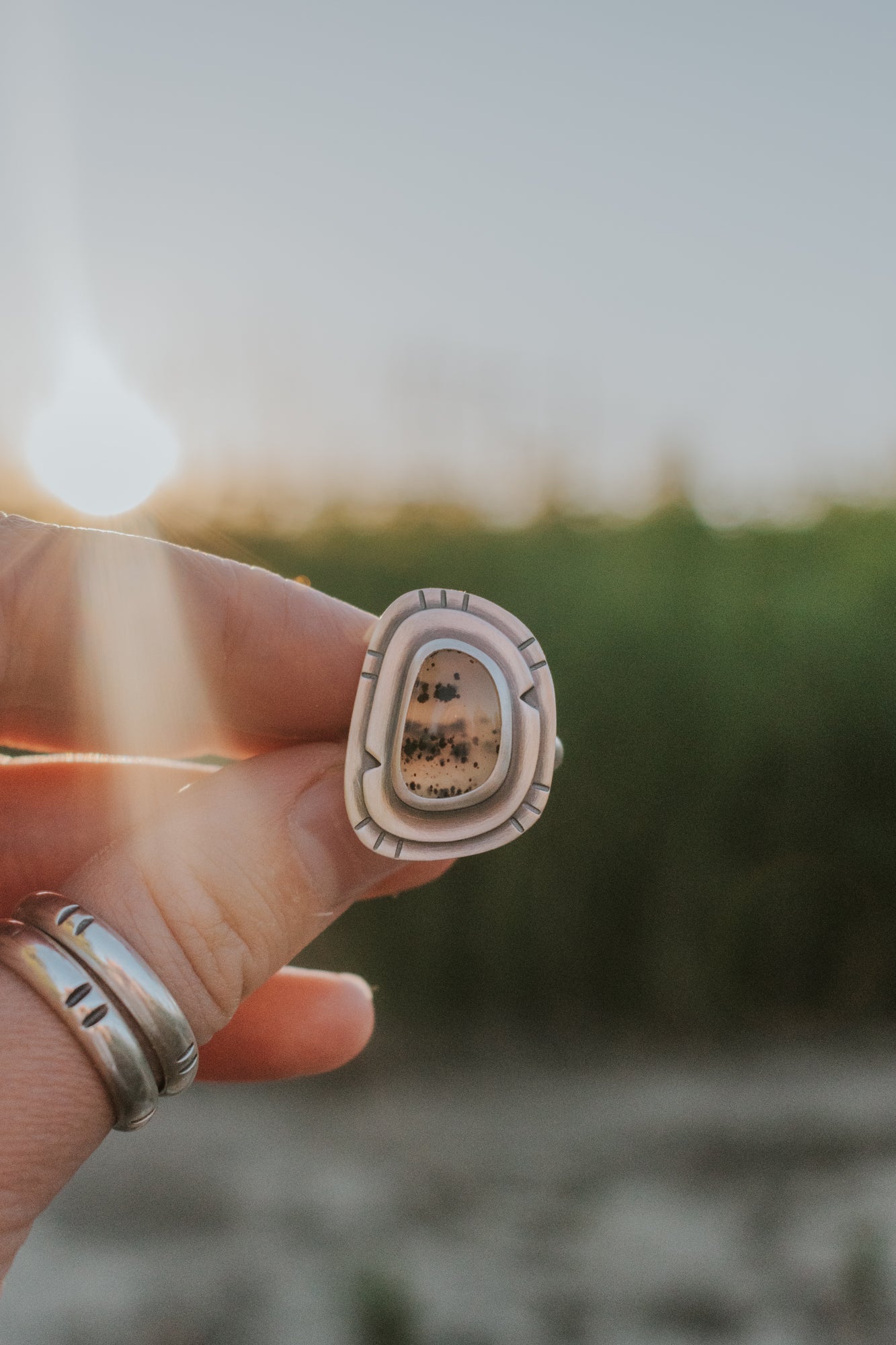A Montana Agate and Sterling Silver ring is  being held up by a hand, with the sun shining over a grassy field in the background. 