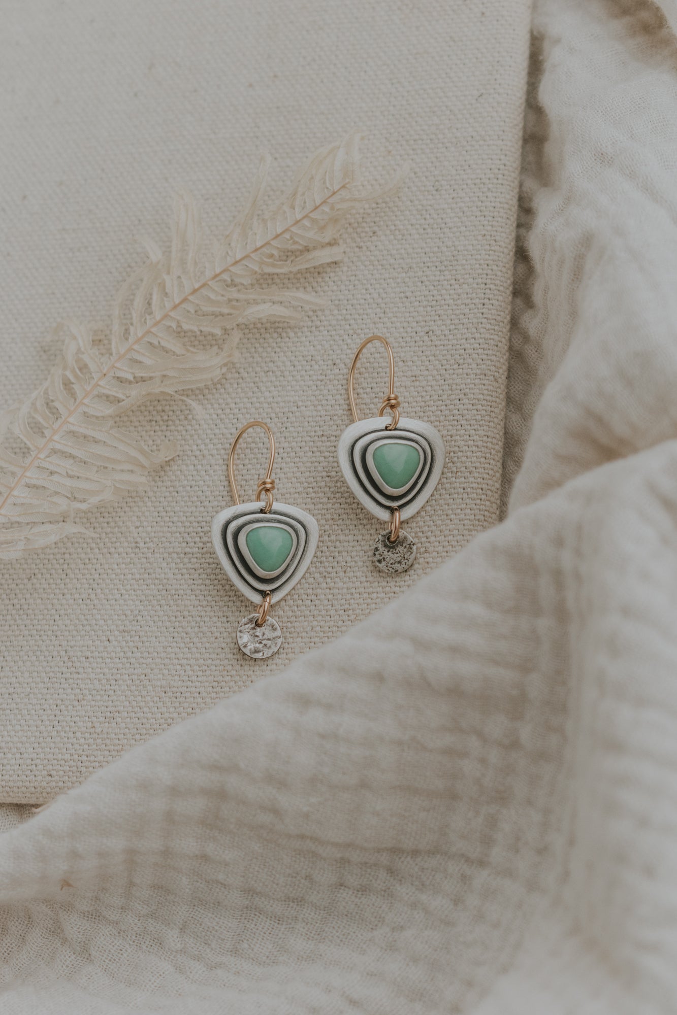 A pair of handcrafted earrings with triangular variscite stones, gold-filled earwires, and silver dangling jump rings, displayed on a textured fabric.